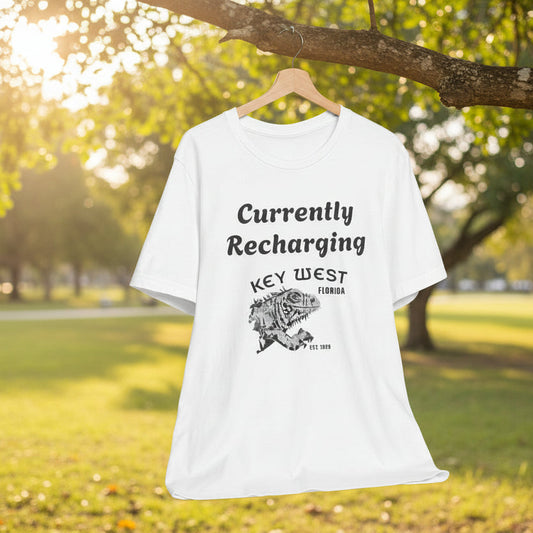 White t-shirt with 'Currently Recharging Key West Florida' text and graphic on a hanger against a park background.