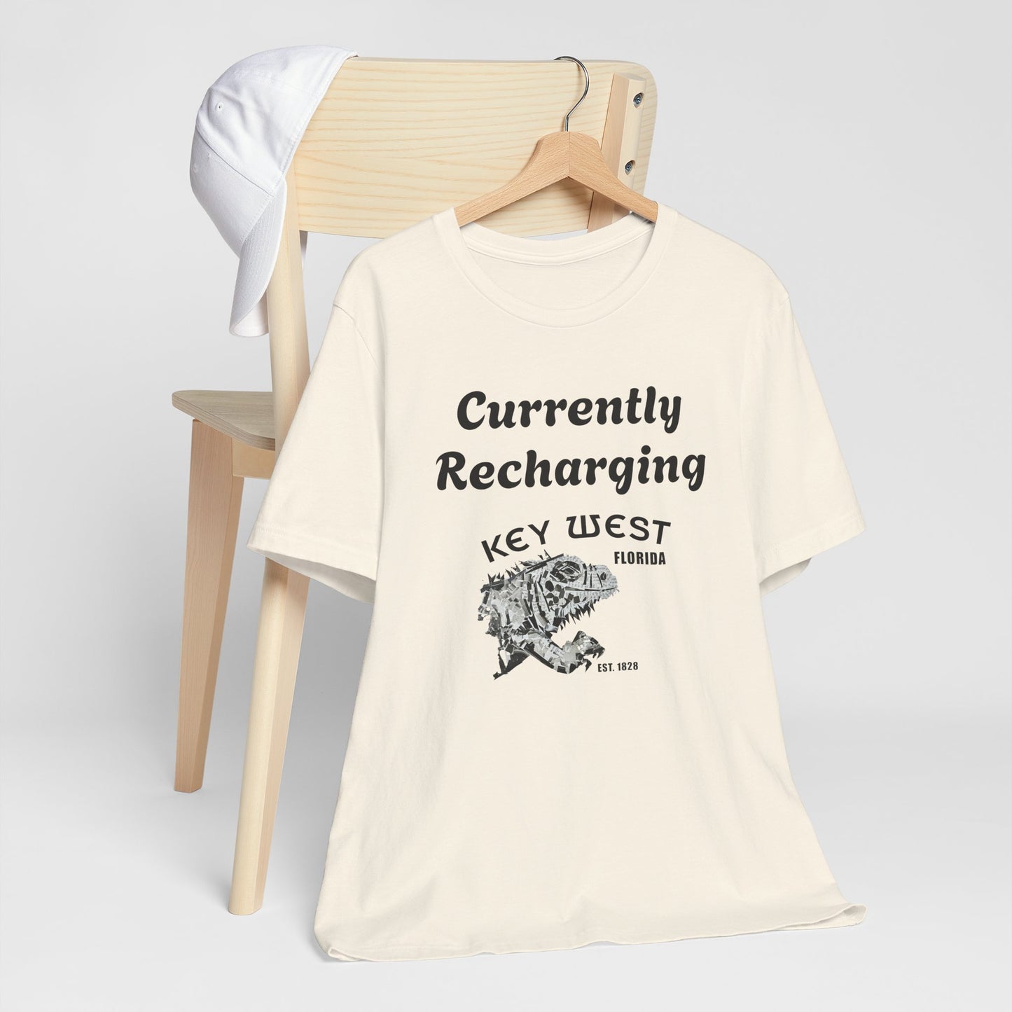 Beige t-shirt with 'Currently Recharging Key West Florida' text and graphic on a wooden chair.