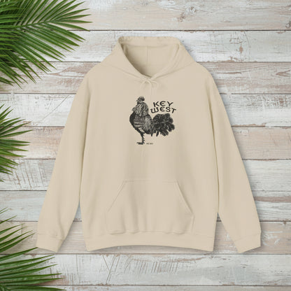 Beige hoodie with rooster graphic and 'Key West' text on a wooden surface with green leaves.