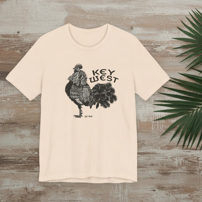 Beige t-shirt with rooster graphic and 'Key West' text on a wooden surface with a palm leaf.