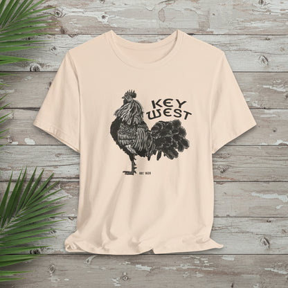 Beige t-shirt with rooster graphic and 'Key West' text on a wooden surface with palm leaves.