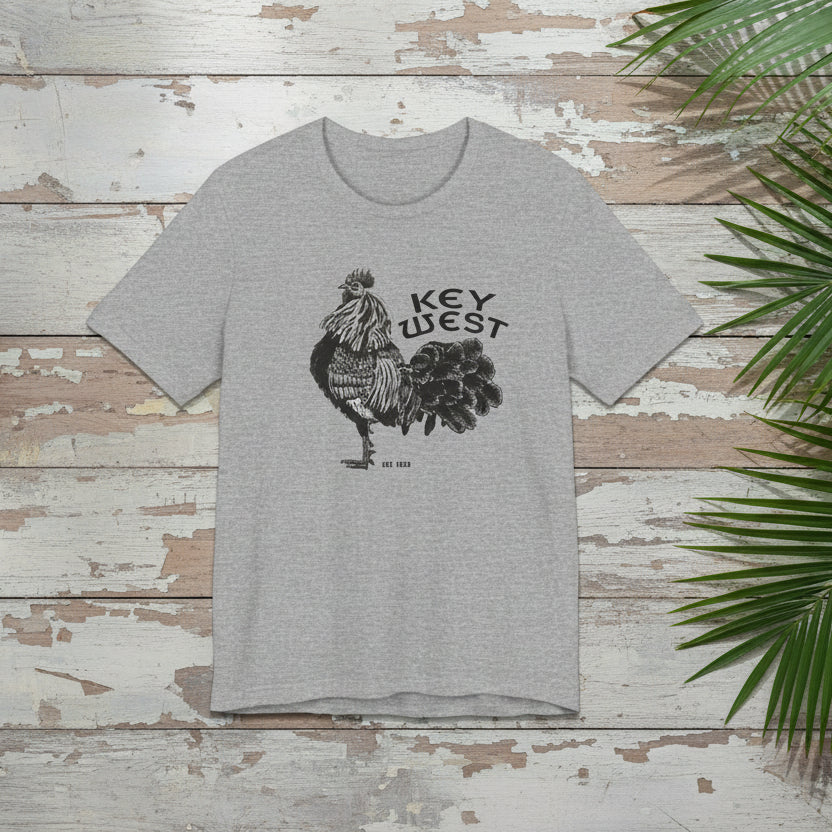 Gray t-shirt with rooster graphic and 'Key West' text on a wooden surface with green leaves.