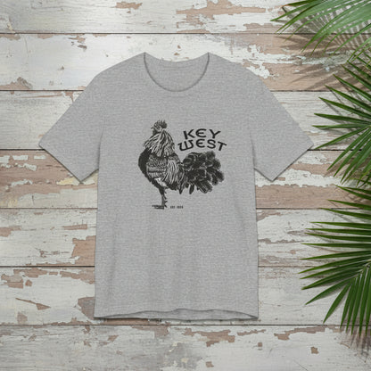 Gray t-shirt with rooster graphic and 'Key West' text on a wooden surface with green leaves.