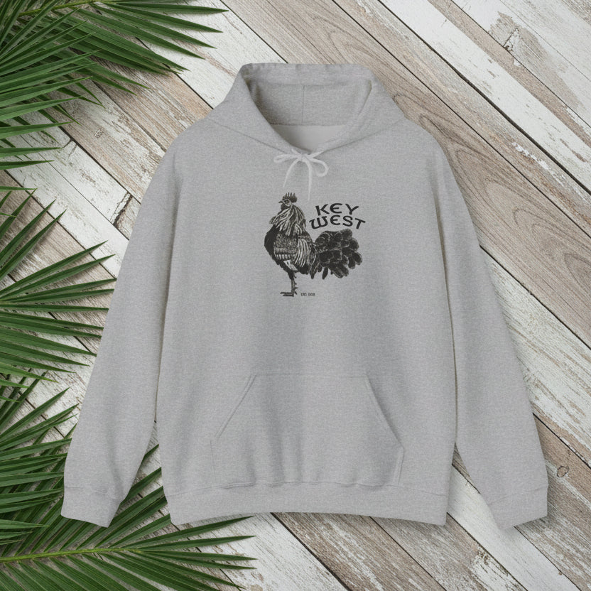 Gray hoodie with rooster graphic and 'Key West' text on a wooden surface with palm leaves.