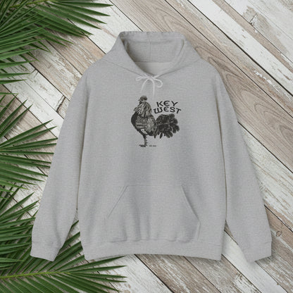 Gray hoodie with rooster graphic and 'Key West' text on a wooden surface with palm leaves.