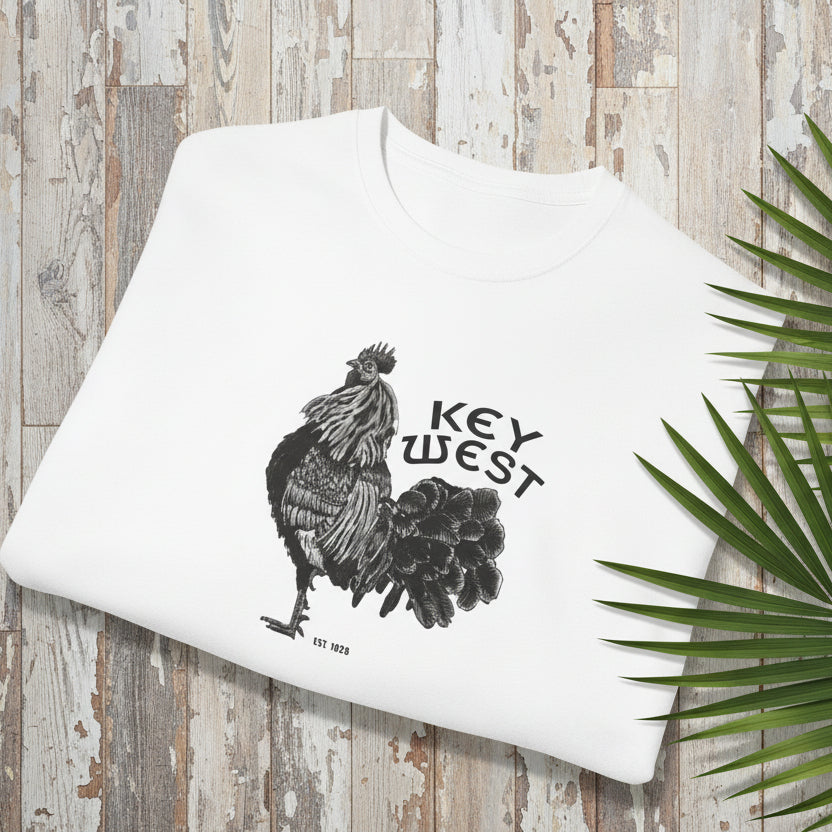 White pillow with rooster design and 'Key West' text on a wooden surface with a palm leaf.