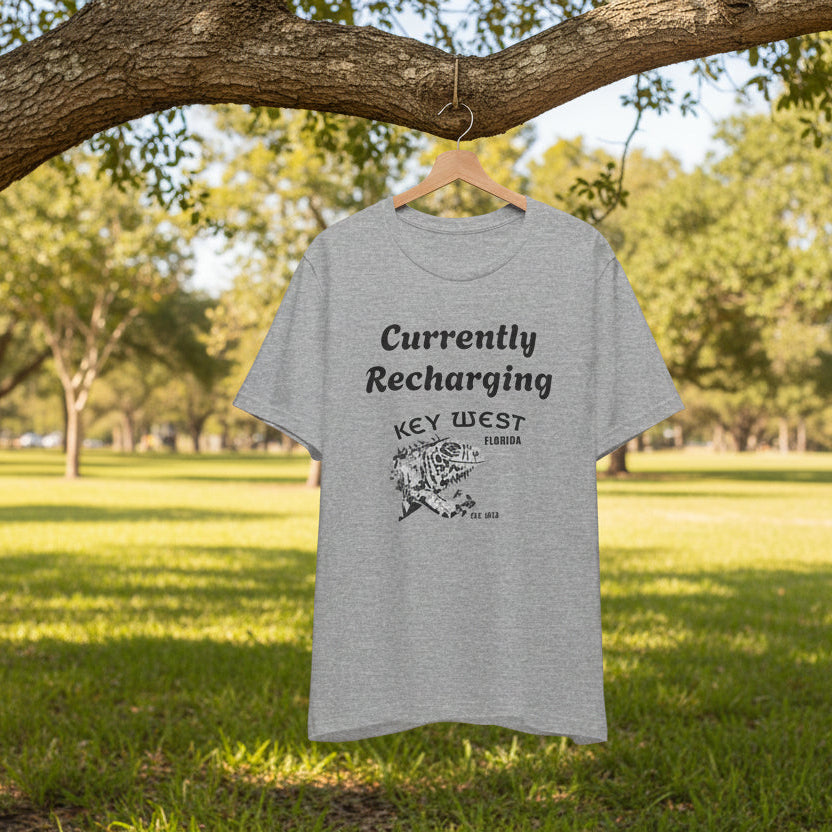 Gray t-shirt with 'Currently Recharging Key West' text and graphic on a hanger under a tree in a park.