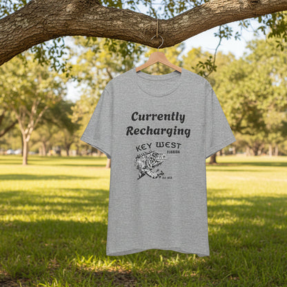 Gray t-shirt with 'Currently Recharging Key West' text and graphic on a hanger under a tree in a park.