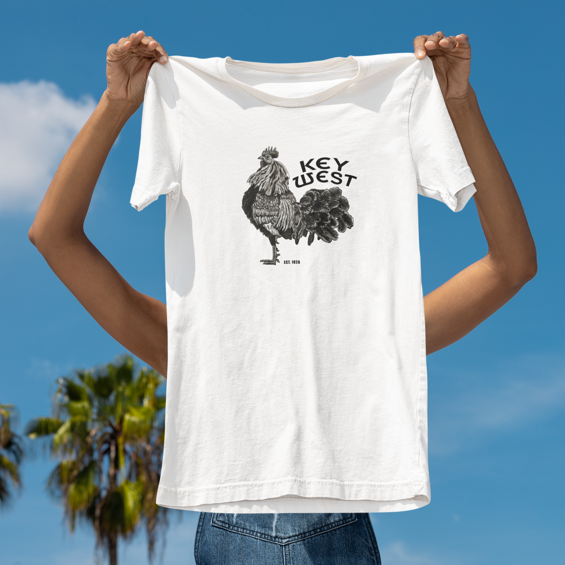 Person holding a white t-shirt with a rooster graphic and 'Key West' text against a blue sky.