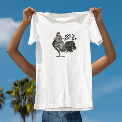Person holding a white t-shirt with a rooster graphic and 'Key West' text against a blue sky.