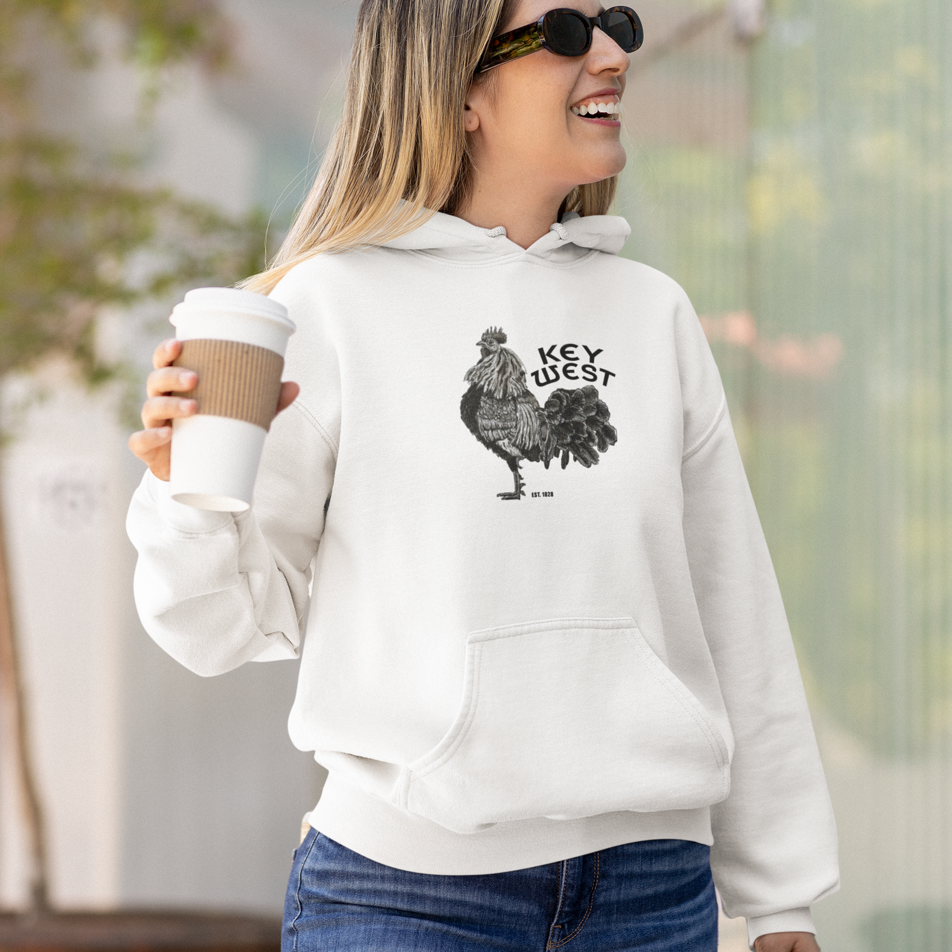 Woman wearing a white hoodie with a graphic design, holding a coffee cup outdoors.